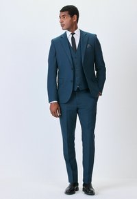 Námořnický modrý třídílný oblek se střihem, který zahrnuje klopu s notchem, pět knoflíků a postranní kapsy. Černé boty dokončují vzhled.