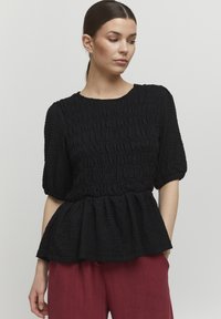 Sort tekstureret bluse med korte pufærmer og peplum-søm. Har rund halsudskæring og detaljer med sammensamlet stof. Bæres med røde bukser.