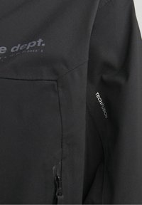 Chaqueta negra con un acabado suave, que presenta un bolsillo con cremallera y texto impreso. Incluye una etiqueta "TECH FUSION" en la manga.