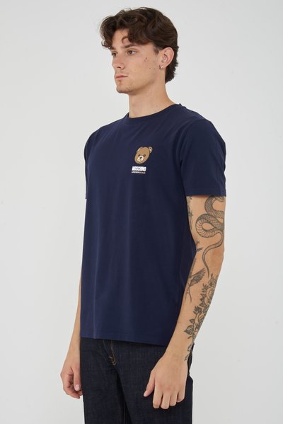 Young man in navy blue Moschino T-shirt with bear logo, showing tattooed right arm, standing against plain background.