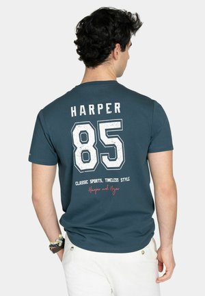 Hombre con camiseta de color verde azulado oscuro con el texto "HARPER 85" y "Classic Sports, Timeless Style" en la espalda, combinada con pantalones blancos.