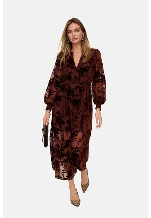 Frau geht, trägt ein durchscheinendes dunkelbraunes Blumenkleid mit Puffärmeln, hält eine dunkle Clutch und trägt spitze beigefarbene Schuhe.