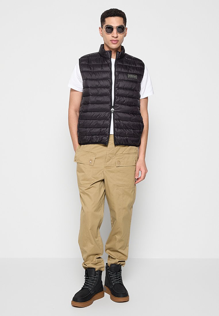 Schwarze, wattierte Weste mit Reißverschluss, über einem weißen T-Shirt getragen. Beigefarbene Cargo-Hosen und schwarze Stiefel vervollständigen das Outfit. Runde Sonnenbrillen setzen ein zusätzliches Detail.