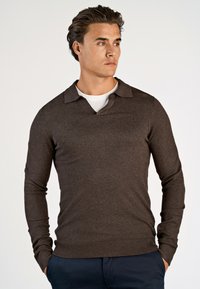 Ung man med mörkt hår som bär en brun långärmad stickad polo över en vit skjorta, med händerna i fickorna på navybyxor, tittar åt sidan.