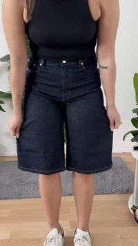 Persona che indossa pantaloni corti in denim scuri a vita alta, all'altezza del ginocchio, e sneakers bianche, che si trova all'interno su un pavimento in legno.