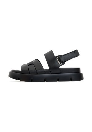 Sandalias negras con un diseño de tiras anchas, hechas de cuero suave. Presentan una suela plana con un borde texturizado y una correa con hebilla ajustable.