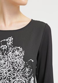 Detailní záběr osoby v černém dlouhém rukávu s bílým složitým květinovým korálkovým vzorem na hrudi.