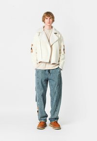 Chaqueta corta de crema con cuello de borrego, jeans holgados azules y zapatos marrones. La chaqueta presenta bordados en las mangas; los jeans tienen cremalleras laterales.