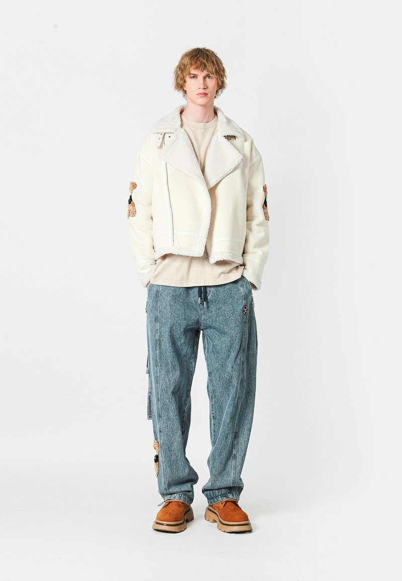 Chaqueta corta de crema con cuello de borrego, jeans holgados azules y zapatos marrones. La chaqueta presenta bordados en las mangas; los jeans tienen cremalleras laterales.