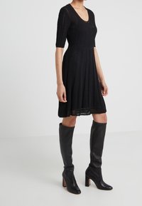 Abito in maglia nera con maniche corte, scollo rotondo, finitura testurizzata e gonna svasata, abbinato a stivali neri aderenti al ginocchio.