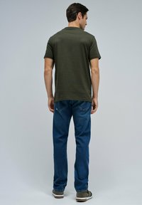 Hombre de pie de espaldas, vistiendo una camiseta verde oliva, jeans azules y zapatillas grises, contra un fondo gris liso.