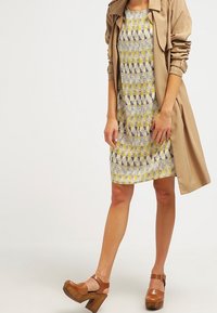 Abrigo beige sobre un vestido de largo hasta la rodilla con un patrón que presenta tonos amarillos, negros y grises. Unas sandalias de plataforma marrones completan el conjunto.