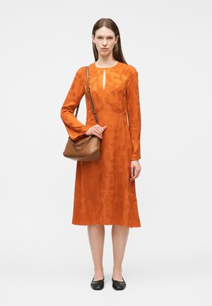 Γυναίκα με πορτοκαλί μίντι φόρεμα με μακριά μανίκια και διακριτικό μοτίβο, κρατώντας καφέ δερμάτινη τσάντα ώμου και φοράει μαύρες μπαλαρίνες μπροστά σε απλό φόντο.