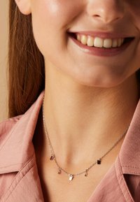 Zilveren ketting met kleine rechthoekige hangers en ronde accenten, gedragen om een gebruinde nek, met een roze overhemd op de achtergrond.