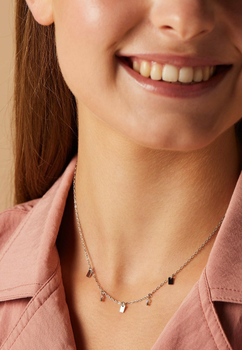 Zilveren ketting met kleine rechthoekige hangers en ronde accenten, gedragen om een gebruinde nek, met een roze overhemd op de achtergrond.