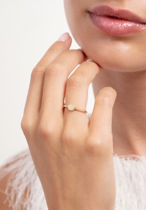 Nahaufnahme einer Hand nahe am Gesicht, die einen dünnen Goldring mit rundem, strukturiertem Fassungsrand am Mittelfinger trägt.