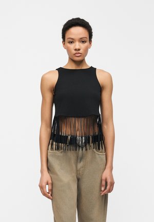 Γυναίκα που φοράει μαύρο αμάνικο κοντό τοπ με φούντες που κρέμονται πάνω από μπεζ παντελόνι ψηλόμεσο και μαύρη ζώνη με ασημένια πόρπη σε σχήμα καρδιάς.