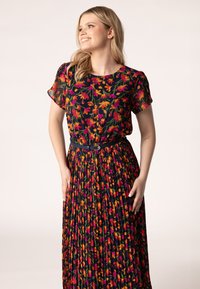 Blumenkleid mit kurzen Ärmeln, schwarzem Grund, lebendigen pinken, orangeroten und grünen Blumen. Faltiger Rock, tailliert mit einem kontrastierenden Gürtel.