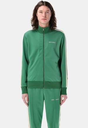 Tuta sportiva verde realizzata in materiale liscio, con strisce laterali bianche e nere. Presenta un colletto alto e un branding minimale su petto e pantaloni.