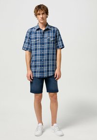 Koszula w kratę z krótkim rękawem w odcieniach niebieskiego z kołnierzykiem, kieszeniami z przodu, zestawiona z dżinsowymi szortami i białymi trampkami. Gładka faktura całości.