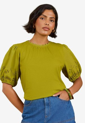 Vrouw met donker haar die een groene blouse met pofmouwen en uitsnijdingen draagt en blauwe jeans, één hand in de zak, draagt creolen en een armband.