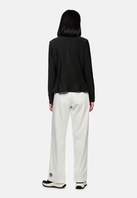 Schwarze Jacke mit einem Peplum-Saum und langen Ärmeln, kombiniert mit lockeren weißen Hosen und kontrastierenden schwarzen Schuhen.