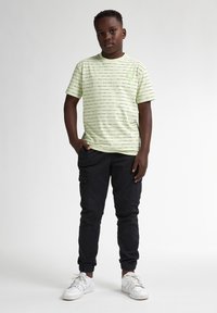 Camiseta de manga corta verde claro con líneas horizontales negras y un pequeño logo, combinada con pantalones cargo negros y zapatillas blancas.