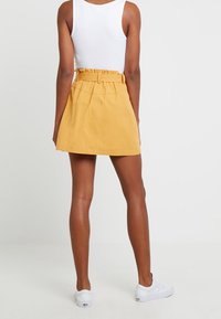 Minigonna di cotone gialla con vita arricciata e cintura, caratterizzata da una texture liscia e un design semplice. Indossata con una maglietta bianca e sneakers.