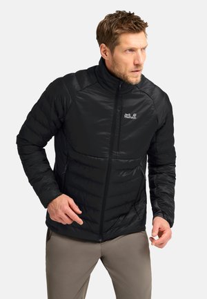 Mann trägt eine schwarze Jack Wolfskin Steppjacke und beige Hose, steht mit konzentriertem Gesichtsausdruck und leicht gebeugten Armen.