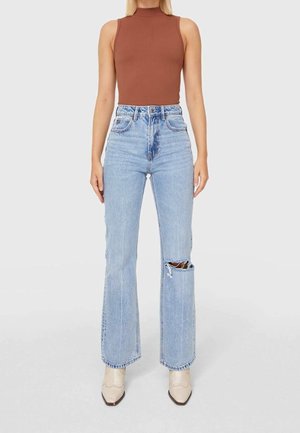 Top marrón de canalé sin mangas combinado con jeans acampanados de talle alto en azul claro que presentan un gran desgarrón en la pierna izquierda.