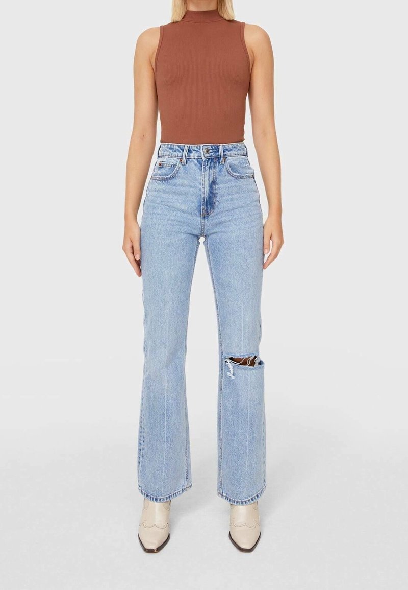 Hnědý žebrovaný top bez rukávů v kombinaci se světle modrými džínami s vysokým pasem a rozšířenými nohavicemi, které mají velký roztrhnutý detail na levé noze.