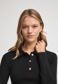 Polo nero a maniche lunghe con una placca con bottoni caratterizzata da bottoni dorati e colletto. Il tessuto sembra liscio e aderente, con dettagli ricamati sulla manica.