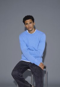 Világoskék pulóver bordás mandzsetta- és nyakkialakítással; fehér ing fölé öltözve, sötétszürke farmernadrággal párosítva. A modell egy fém széken ül.