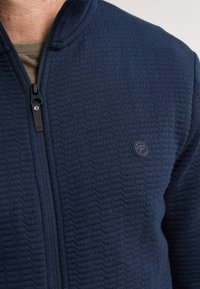 Námořnická modrá strukturovaná zipová bunda s vysokým límcem, má jemný vzor ve tvaru cik-cak a kulatou nášivku na levé straně.