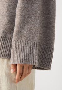 Détail d'un pull en maille marron clair avec des poignets côtelés. Texture douce, coupe légèrement ample, présentant un ourlet droit et des coutures minimales.