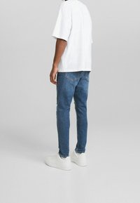 Biały t-shirt z krótkim rękawem, niebieskie dżinsy z lekkimi przetarciami oraz białe sneakersy, widoczne od tyłu, na neutralnym tle.