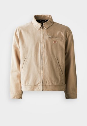 Beige canvas jacket featuring a zip front, stand collar, and two side pockets. Includes a small logo patch on the chest.