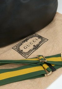 Sac en cuir noir posé sur une housse anti-poussière beige Gucci avec une sangle en tissu rayée verte et jaune et une quincaillerie dorée.