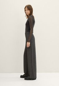 Haut gris à manches longues en tissu texturé et scintillant, rentré dans un pantalon large taille haute de couleur sombre. Le modèle porte des chaussures à plateforme noires et chunky.