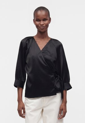 Personne portant un chemisier cache-cœur en satin noir à manches trois-quarts et un pantalon blanc, debout devant un fond blanc uni.