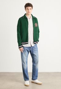 Groene gebreide cardigan met paarse kraagaccenten, geborduurd logo, gecombineerd met gestreept shirt en lichtblauwe jeans. Witte sneakers maken de outfit compleet.