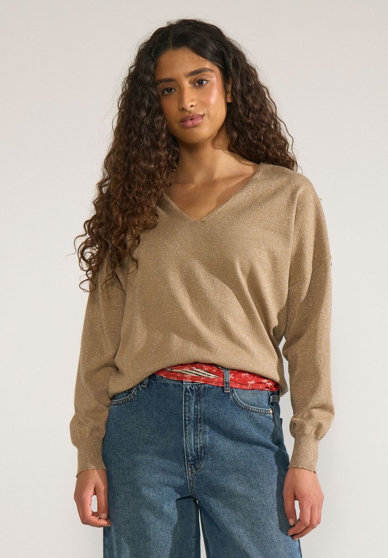 Femme aux longs cheveux bouclés portant un pull beige scintillant à col en V et un jean bleu taille haute avec une ceinture rouge à motif.