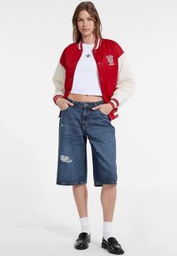 Rode en crèmekleurige varsityjas met geribde manchetten en logo, gecombineerd met blauwe denim Bermuda-short met verweerde details. Zwarte loafers en witte sokken.