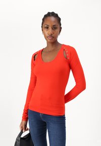 Rotes Langarmshirt mit ausgeschnittenen Schultern und metallischen Ösen. Hat ein körperbetontes Design und eine weiche Textur, kombiniert mit blauen Jeans.
