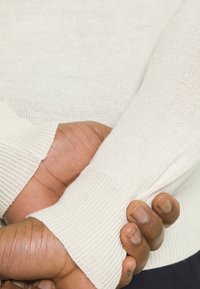 Ljus beige stickad tröja med ribbade mudd, tillverkad av mjukt material och har en texturerad yta. Händerna är hopknäppta nära ärmen.