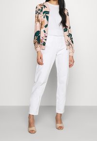 Veste bomber florale rose avec des accents verts et bordeaux, assortie à un pantalon blanc et des talons ouverts couleur nude.