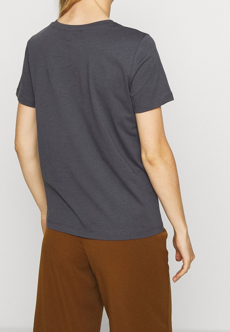 Personne portant un t-shirt uni gris foncé à manches courtes et un pantalon marron ample, debout, dos à la caméra.