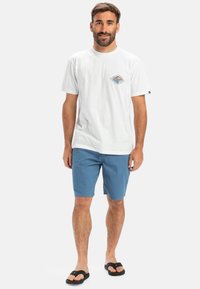 Hvid bomulds t-shirt med et grafisk design på venstre bryst, parret med blå shorts og sorte flip-flops. Står på en neutral baggrund.