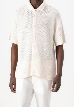 Uomo che indossa una camicia a maniche corte color crema in maglia a rete con colletto e bottoni, abbinata a pantaloni bianchi, in piedi davanti a uno sfondo bianco.