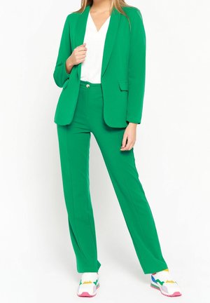 Frau trägt grünen Blazer und Hose mit weißem Top und bunten Sneakers, steht vor weißem Hintergrund.